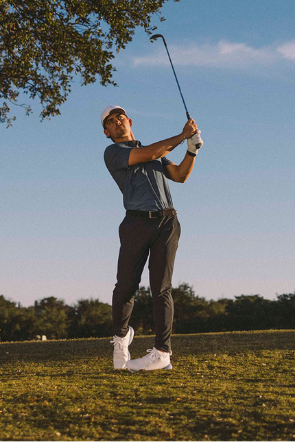 A man in a golf outfit swings a club on a golf course, positioned to the right of a large tree under a clear blue sky.