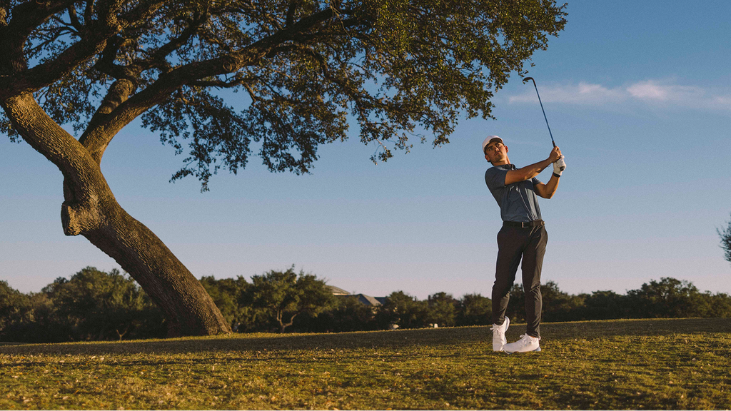 A man in a golf outfit swings a club on a grassy golf course under a clear blue sky, with a large tree on the left and other trees in the background.